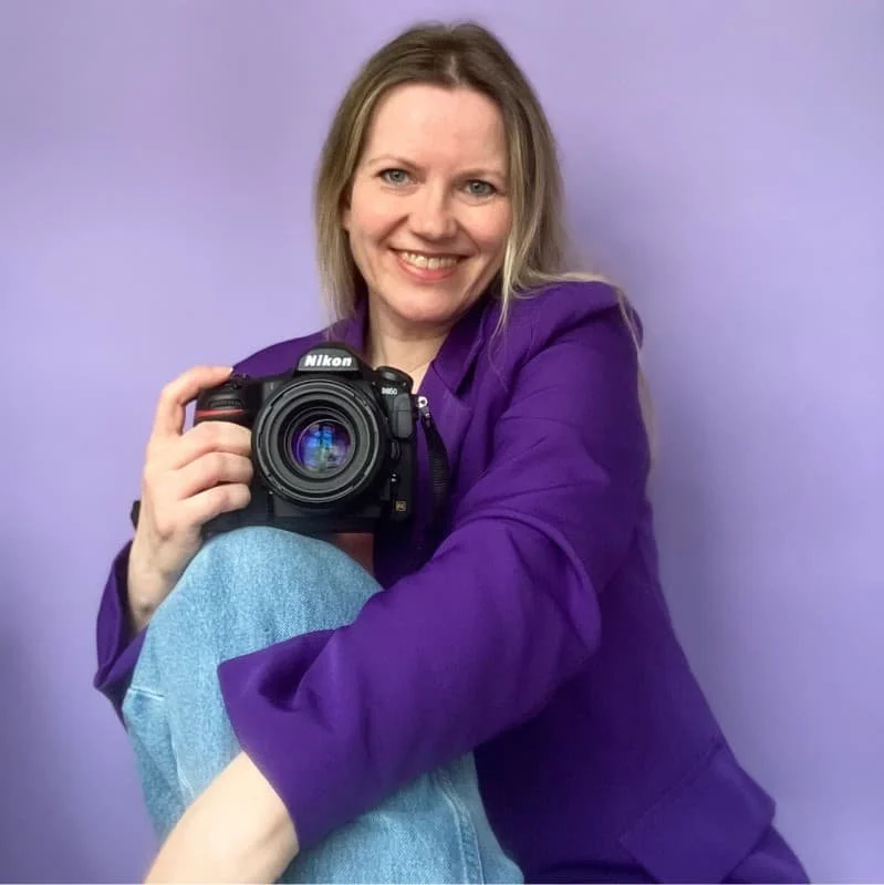 Miriam hat eine Fotokamera in der Hand, traegt einen Lila Blazer und eine Jeans. Ihr Haar ist lang und blond.