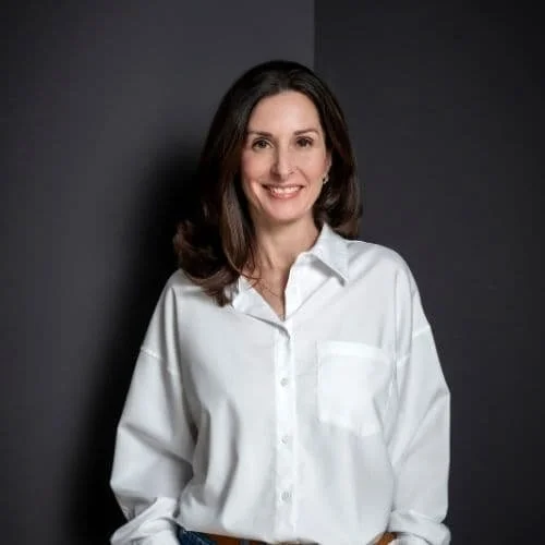 Eine Frau mit braunen Haaren trägt eine weiße Bluse und lächelt, steht vor einer dunklen Wand.