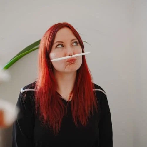 Nicole traegt einen schwarzen Pullover, hat einen Stift zwischen Nase und ihrer Oberlippe, ihr Haar ist rot und lang