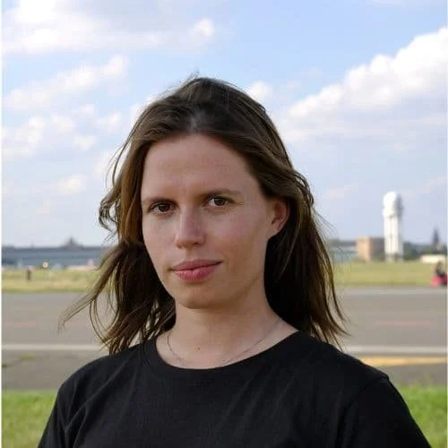Junge Frau mit braunen Haaren vor einer Landschaft mit Himmel, Wiesen und Turm im Hintergrund.