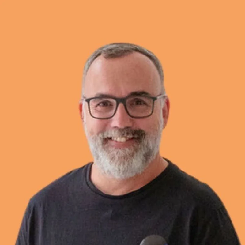 Porträt eines Mannes mit Brille und grauem Bart vor orangem Hintergrund.