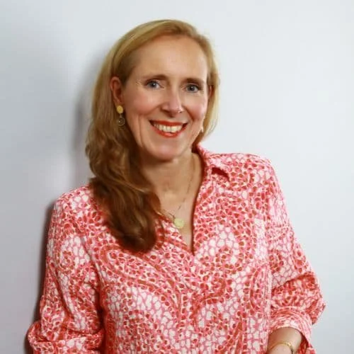 Eine lächelnde Frau mit langen, lockigen, roten Haaren, trägt eine pink-weiße gemusterte Bluse und steht vor einem weißen Hintergrund.