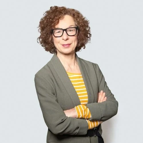 Frau mit lockigem Haar, Brille, grauem Anzug und gelb-weiß gestreiftem Hemd vor weißem Hintergrund.