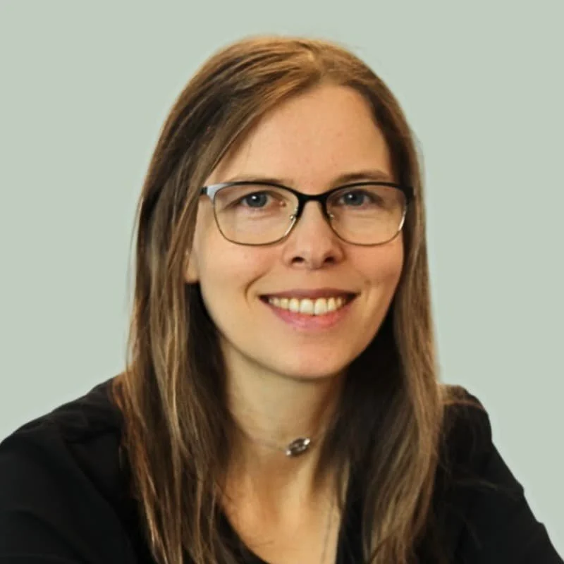 Porträt einer lächelnden Frau mit Brille und langen braunen Haaren vor hellgrünem Hintergrund.