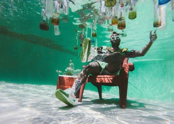 Eine Person sitzt auf einem Stuhl im Wasser, umgeben von hängenden Flaschen und Gläsern, trägt eine bunte Kleidung, Turnschuhe und eine Taucherbrille, dabei lächelnd und ein Daumen nach oben gebend.