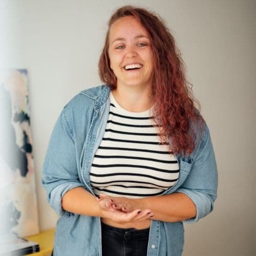 Junge Frau mit lockigem roten Haar, lächelnd, trägt ein gestreiftes T-Shirt und eine Jeansjacke, steht in einem Raum.