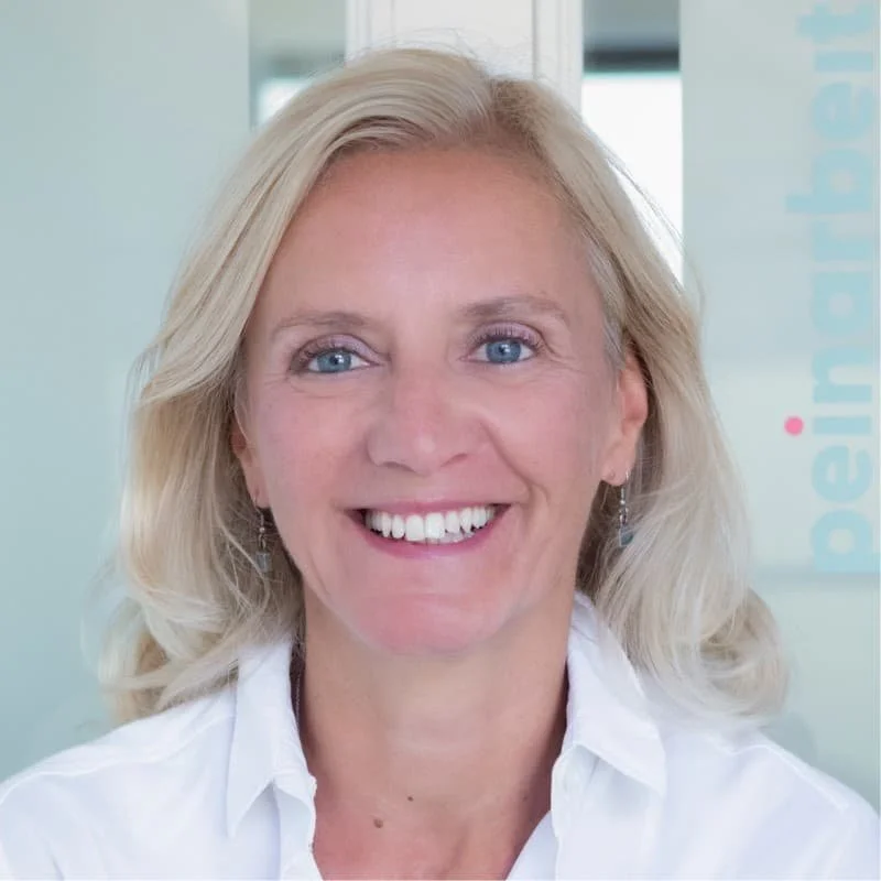 Nahaufnahme einer lachenden Frau mit blonden, lockigen Haaren, trägt weiße Bluse, vor einem Büro-Hintergrund.