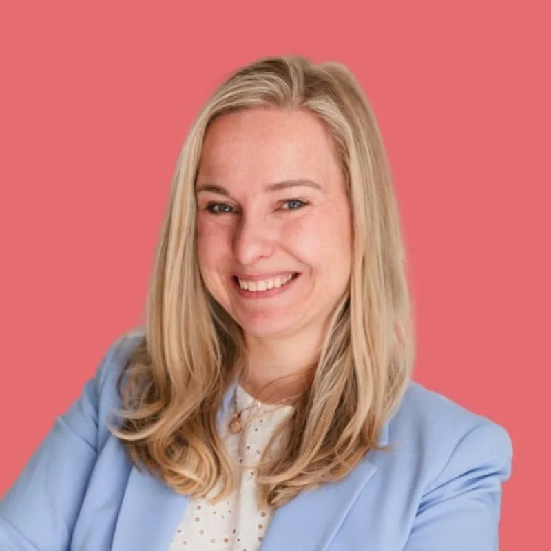 Katrin hat langes blondes Haar, sie traegt einen hellblauen Blazer und ein weisses Shirt. Der Hintergrund des Fotos ist hellrot.