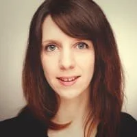 Porträt einer jungen Frau mit braunen, schulterlangen Haaren und einem schwarzen Oberteil, vor hellgrauem Hintergrund.