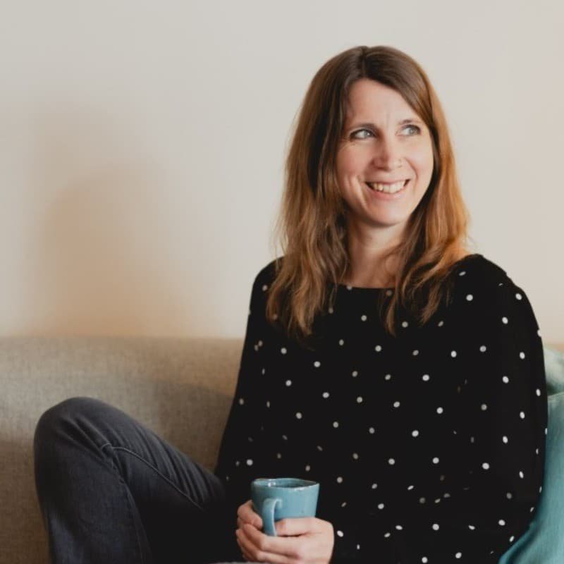 Eine Frau sitzt auf einem Sofa, hält eine Tasse und lächelt. Sie trägt ein schwarzes Oberteil mit weißen Punkten.