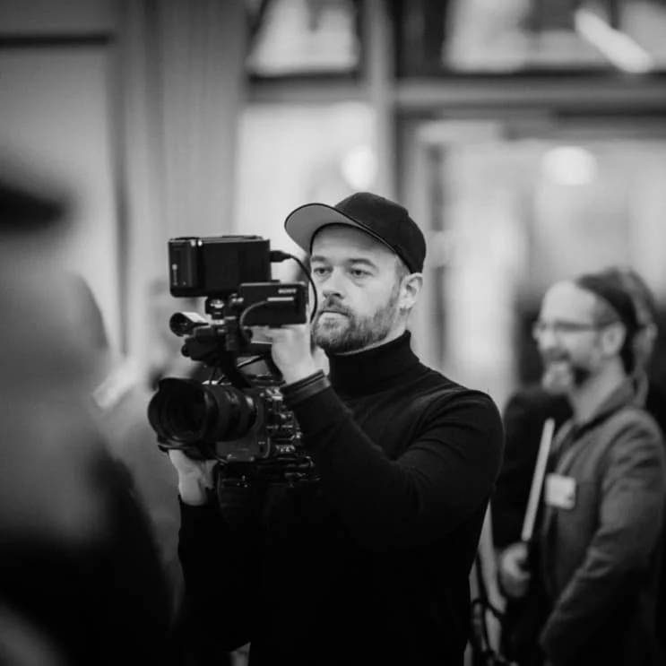 Ein Mann filmt mit einer professionellen Kamera in einem Raum, im Hintergrund sind zwei lachende Frauen.