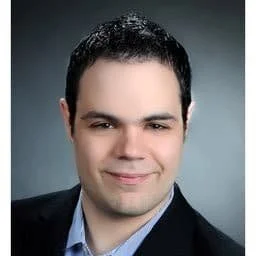 Porträt eines Mannes in formellem Anzug, lächelnd, dunkler Hintergrund.