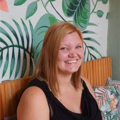 Eine Frau mit rotblondem schulterlangem Haar udnd einem schwarzen T-Shirt. Im hintergrund eine Florale Tapetenwand.