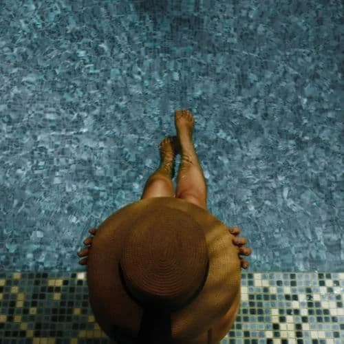Person mit Hut sitzt am Poolrand mit Blick ins Wasser