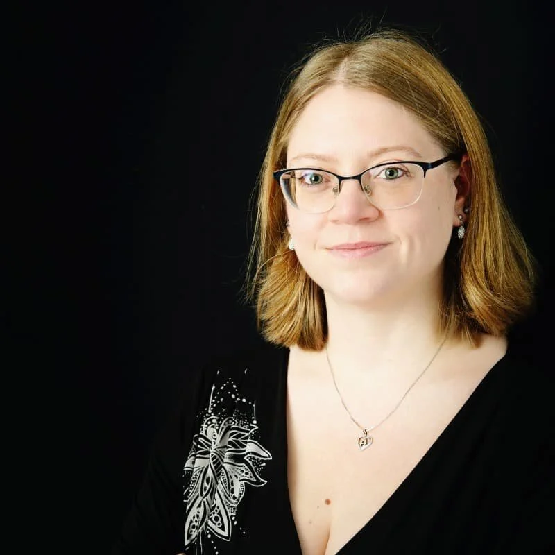 Eine Frau mit roten, hallangen Haaren trägt eine Brille, Kette und ein schwarzes Kleid mit V-Ausschnitt.