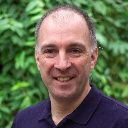 Ein Mann mit kurzen, dunklen Haaren und einem schwarzen Polo-Shirt vor einem grünen Hintergrund.