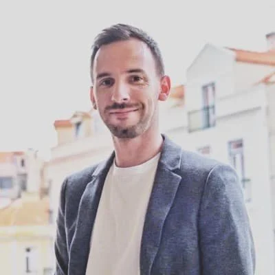 Ein Mann mit dunklem Haar und Bart, trägt ein graues Sakko und ein weißes T-Shirt, steht vor einer Stadt mit mehrstöckigen Gebäuden.