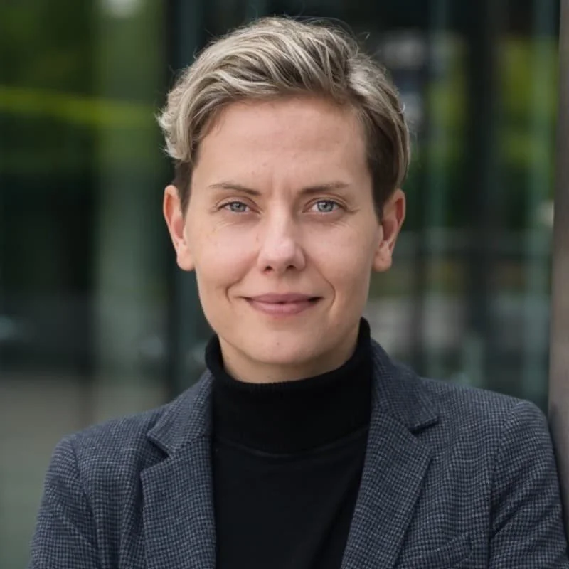 Nahaufnahme einer jungen Frau mit kurzen, blonden Haaren, trägt einen schwarzen Rollkragenpullover und eine graue Blazer, im Hintergrund eine moderne Gebäudefassade