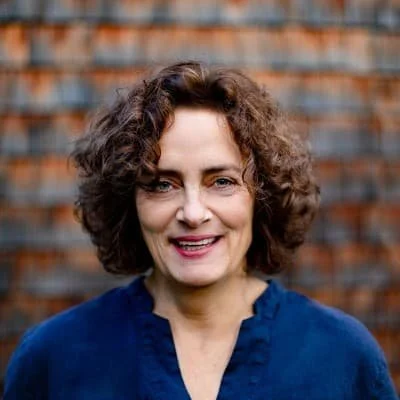 Porträt einer lachenden Frau mit lockigem braunem Haar vor einer Backsteinmauer, trägt eine dunkle Bluse.