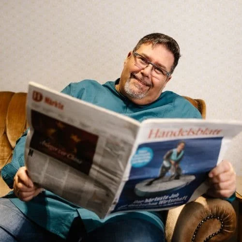 Ein Mann mit Brille und Bart liest eine Tageszeitung in einem Wohnzimmer.