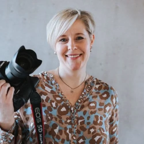 Frau mit kurzen blonden Haaren, lächelnd, hält eine Kamera in der Hand, trägt ein Leopardenmuster-Shirt, vor einem neutralen Hintergrund