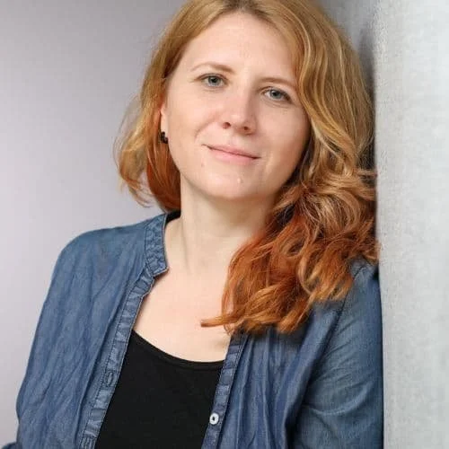 Junge Frau mit lockigem roten Haar, trägt einen blauen Hemd und ein schwarzes Top, lehnt an eine Wand.