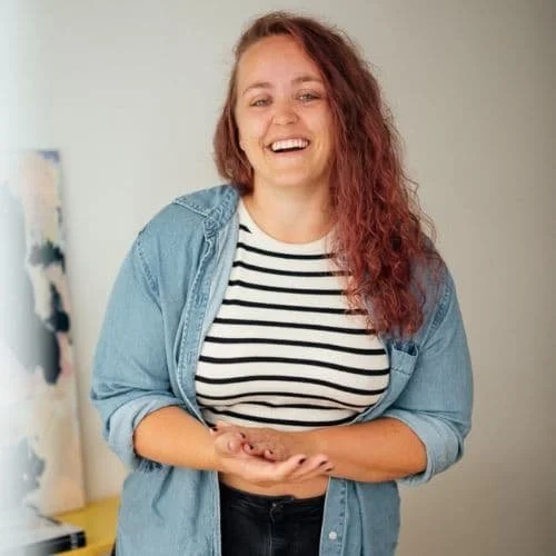 Eine lachende junge Frau mit roten, lockigen Haaren, Kleidung im Stil eines Streifen-Tops und Jeans, steht in einem Raum mit neutralem Hintergrund.