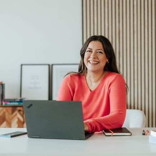 Frau sitzt am Tisch mit Laptop, Notizbuch und Stift, lächelt in einem modernen Büro.