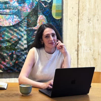 Frau sitzt an einem Holztisch vor einem Laptop, mit einer Tasse links vor sich, im Hintergrund ein abstraktes Wandbild.