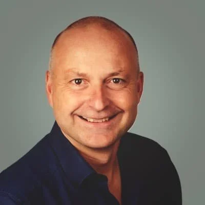 Nahaufnahme eines lächelnden Mannes mit kurzem, blondem Haar, in dunkler Hemd, grauer Hintergrund.