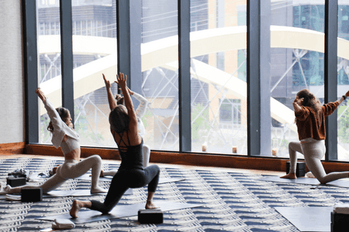 Three women doing yoga in a bright, modern studio with large floor-to-ceiling windows showing a cityscape.