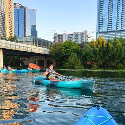 Person kayaking on a river in an urban area with high-rise buildings and greenery in the background.