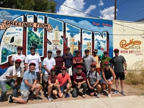Group of young cyclists in front of a vintage mural mural reading 'Greetings from' with cityscapes, castles, and trees, on a sunny day.
