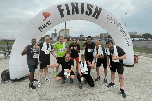 Group of nine runners celebrating at the finish line of a race, standing under a large inflatable arch that reads 'FINISH' with sponsor logos, some giving thumbs up and smiling.