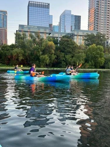 Four people kayaking on a river with city skyscrapers in the background.