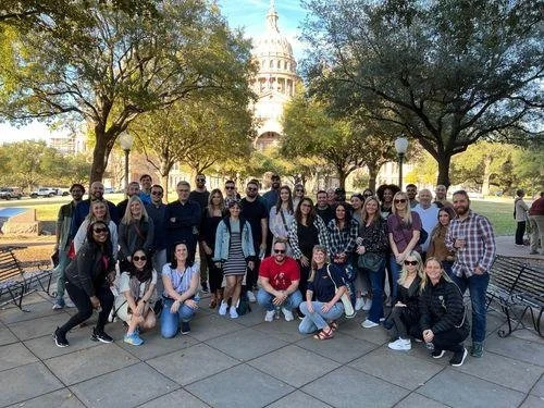 Group of people posing outdoors in front of trees and a government building with a dome, possibly in a park or historic site.