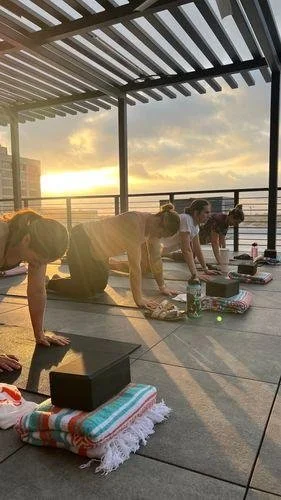Group of people doing yoga on a balcony at sunset with a cityscape in the background.