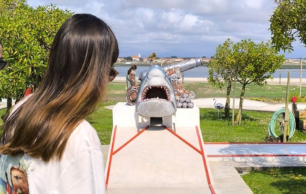 Pessoa observando uma escultura de um tubarão com boca aberta num Minigolfe com árvores, espaço ao ar livre, e uma vista para o rio ao fundo.
