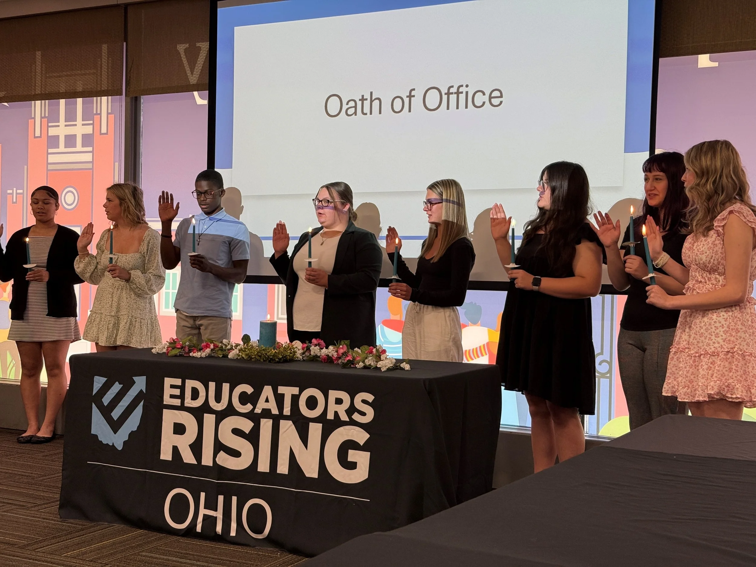 Group of people standing on stage during a ceremony, holding candles, with a table in front that says 'Educators Rising Ohio', and a screen behind displaying 'Oath of Office'.
