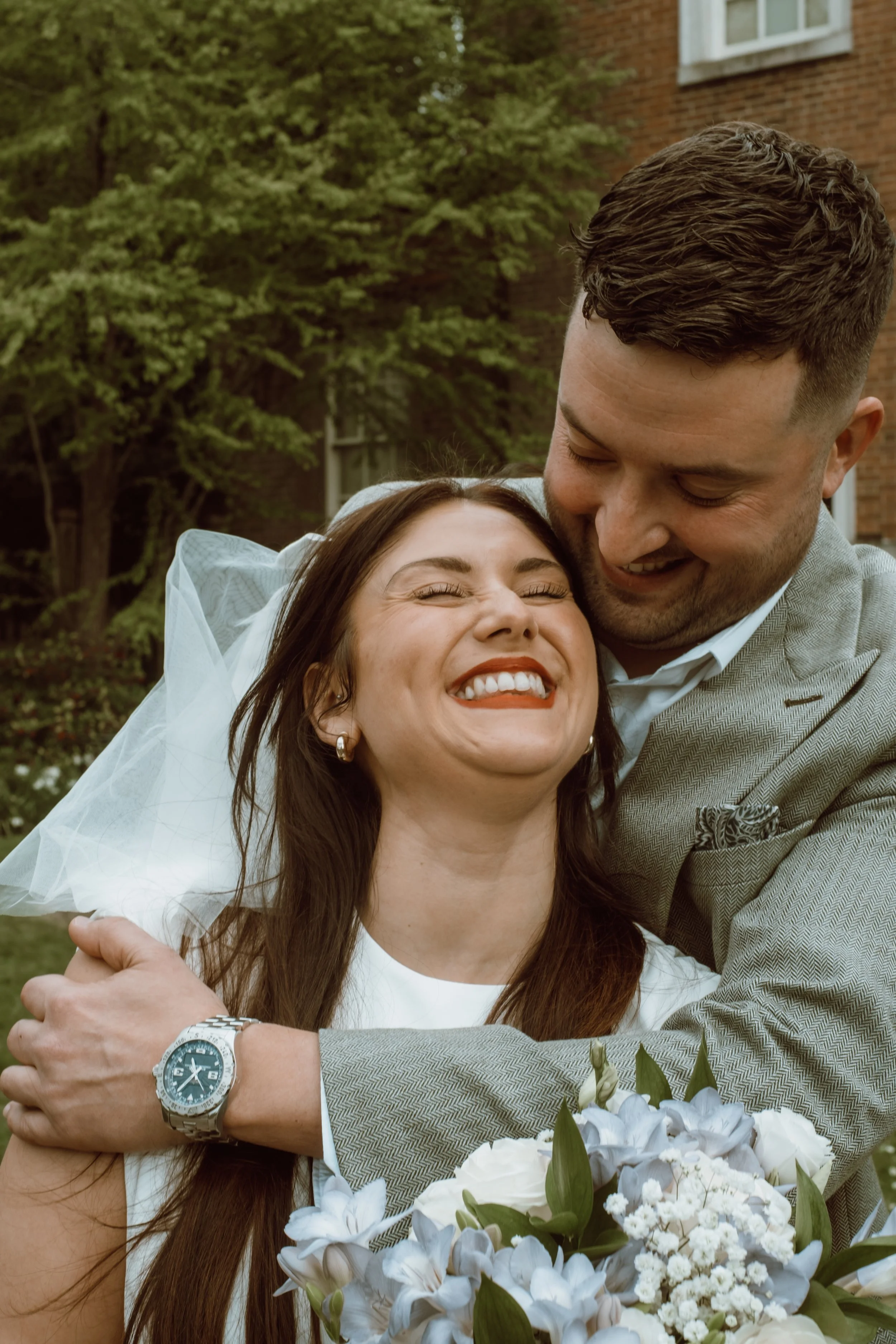 A joyful bride and groom embrace outdoors, smiling and laughing, with flowers and greenery in the background.