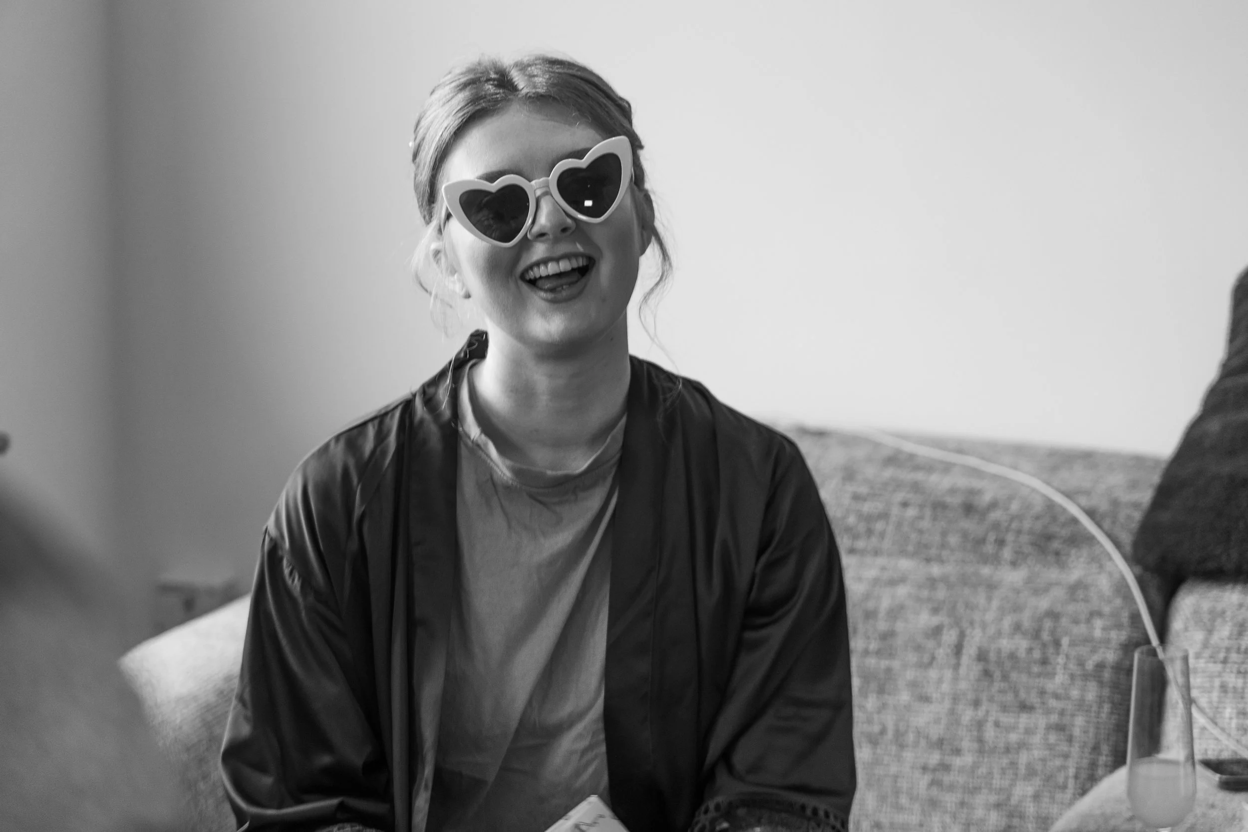 A woman smiling and wearing heart-shaped sunglasses, sitting on a couch with a drink and a remote control in front of her.