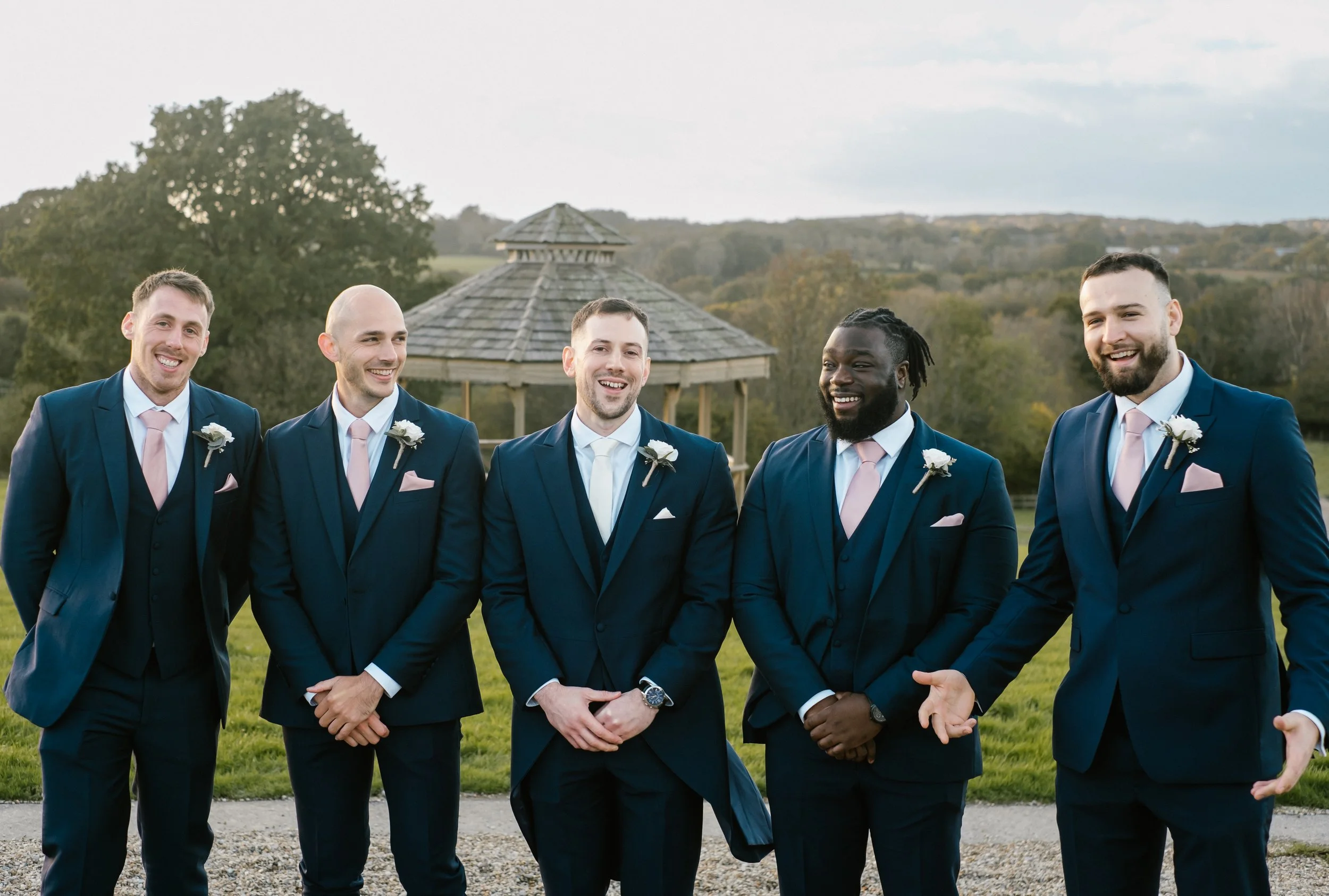 Five men in matching dark blue suits, white shirts, and pink ties standing outdoors, smiling and laughing during a wedding celebration, with a gazebo and trees in the background.