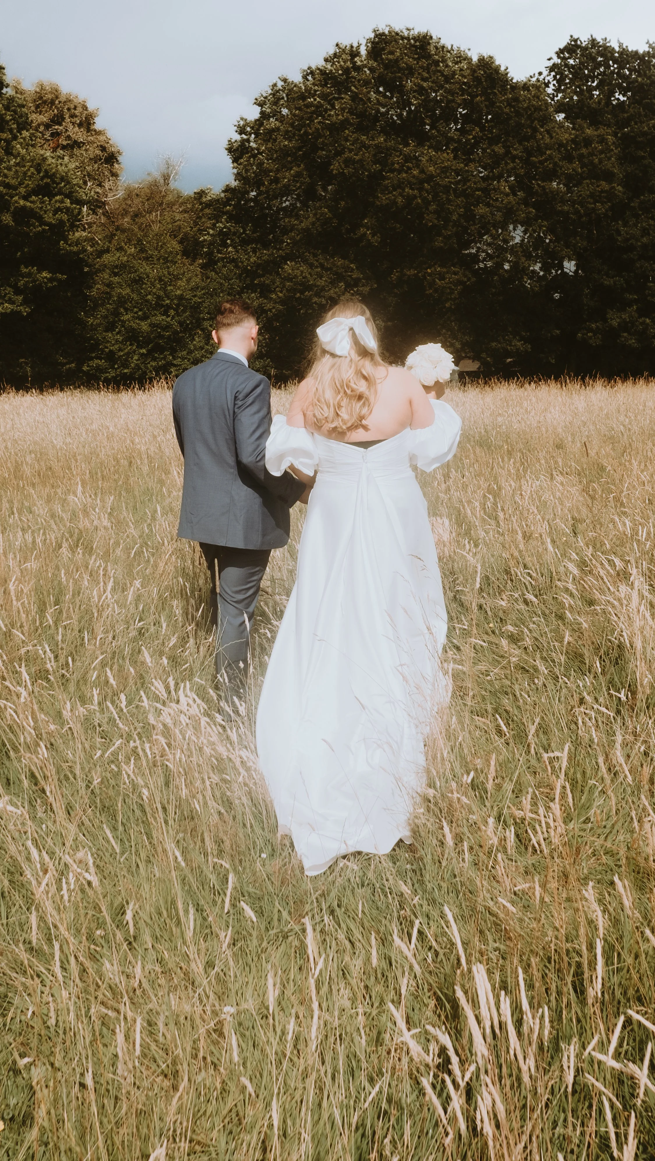 Bride and groom walking through a grassy field with trees in the background.