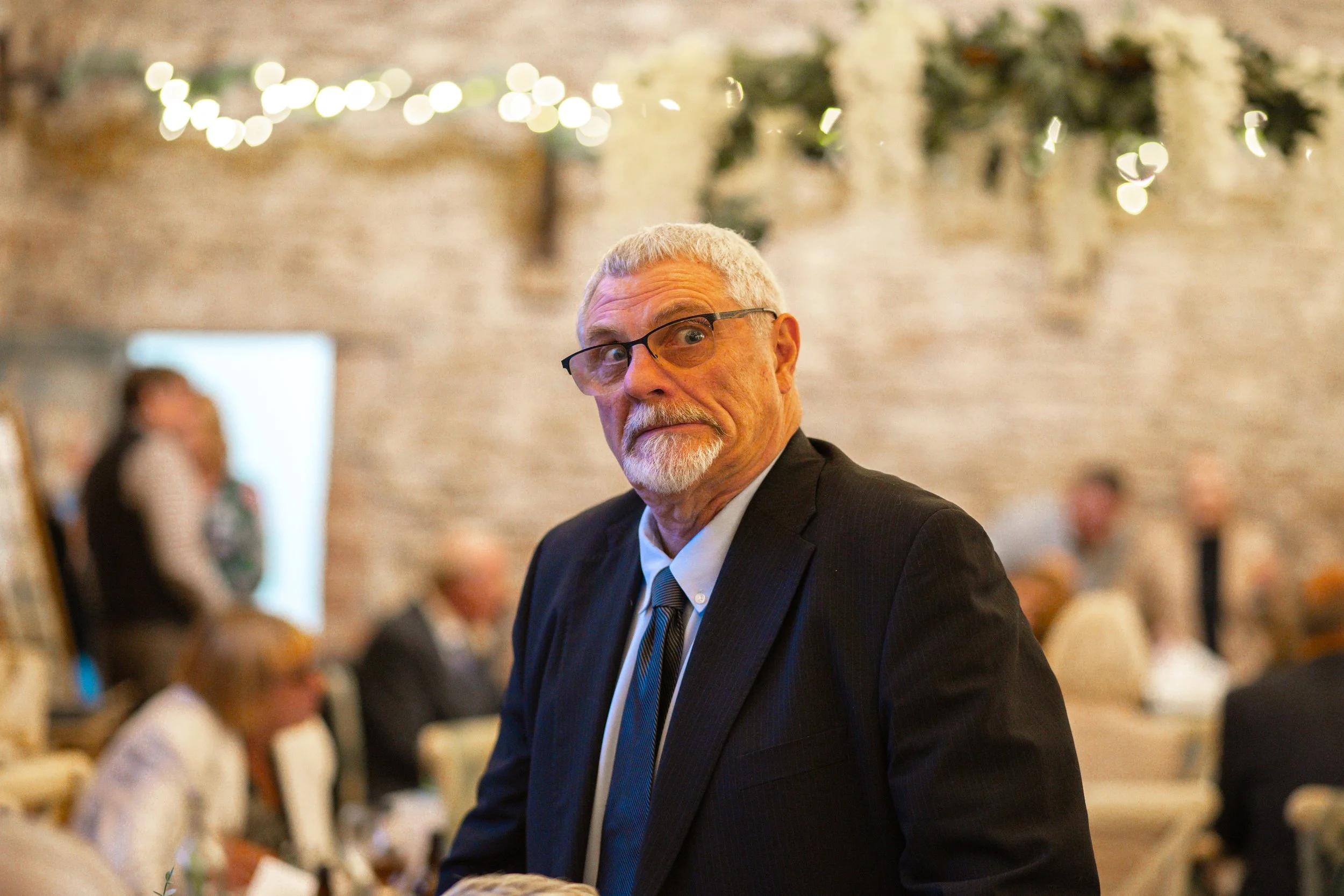 An older man with glasses, gray hair, and a beard wearing a suit, standing at a social event with a brick wall and blurry guests in the background.