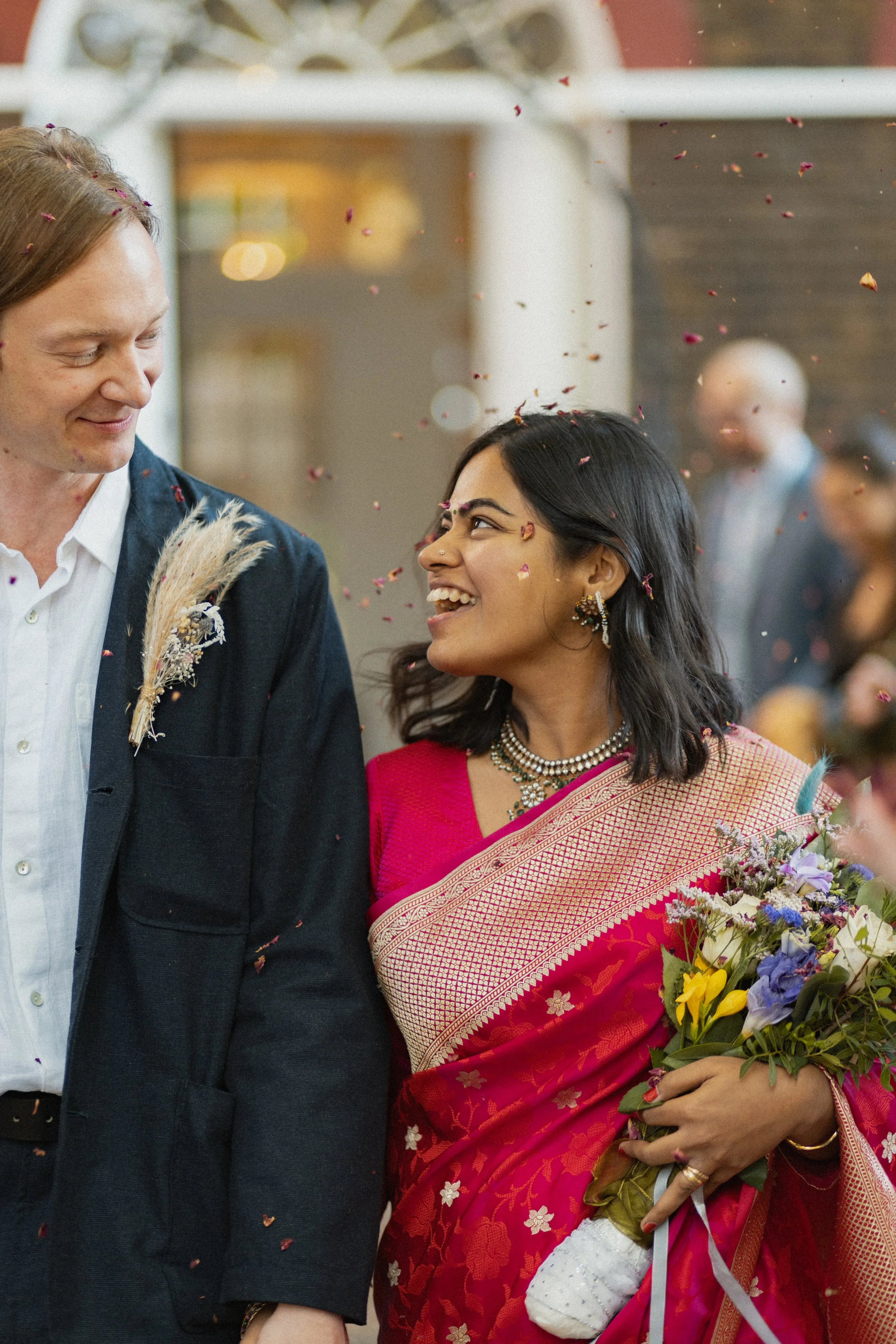 A woman dressed in a traditional pink and gold sari holding a bouquet of flowers, smiling at a man in a dark blazer with a white shirt, at a celebration with confetti in the air.
