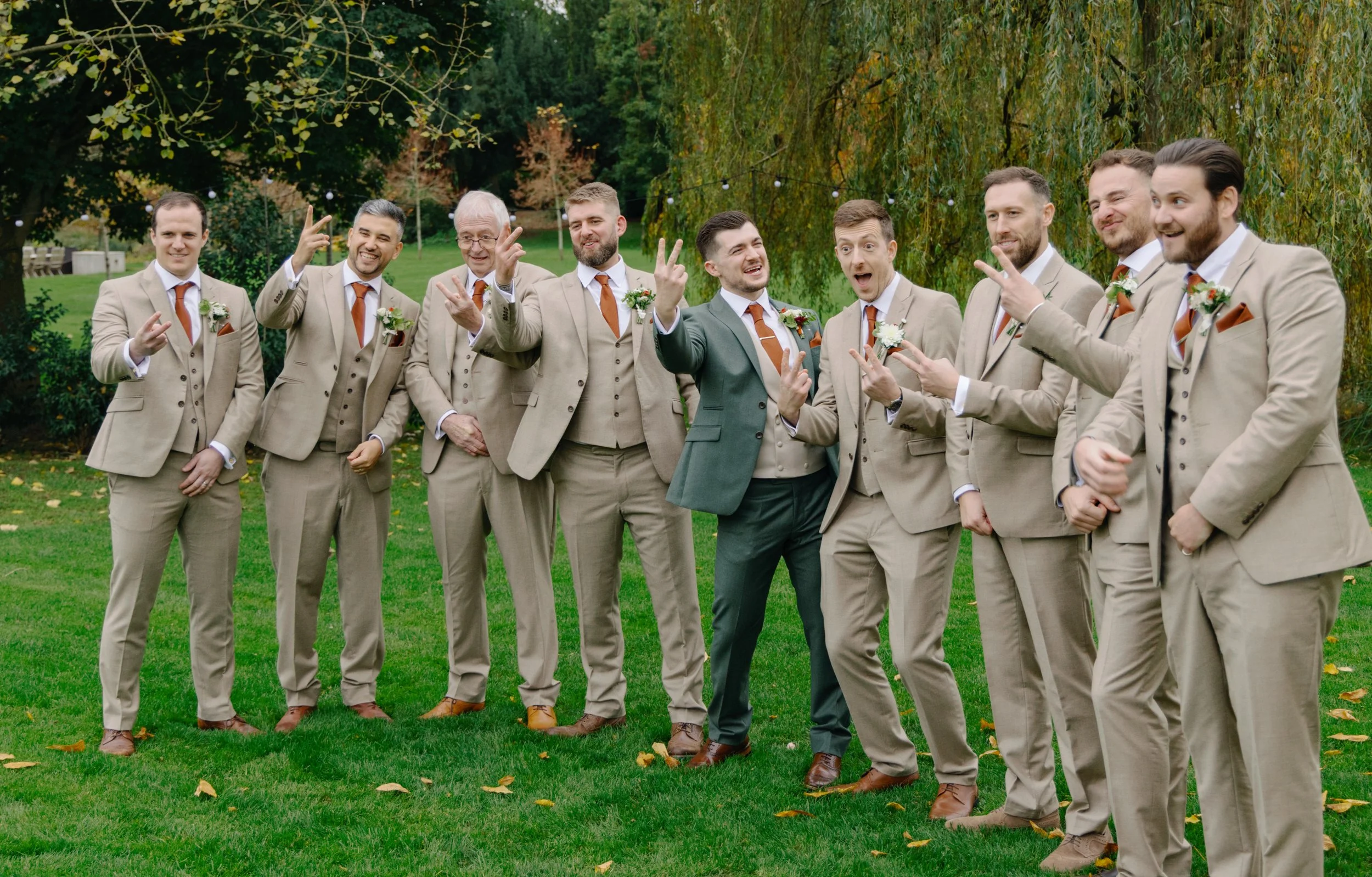 A group of eleven men in beige suits with orange ties and boutonnieres, standing outdoors on green grass with trees in the background. They are smiling, making playful gestures, and appear to be celebrating at a wedding.