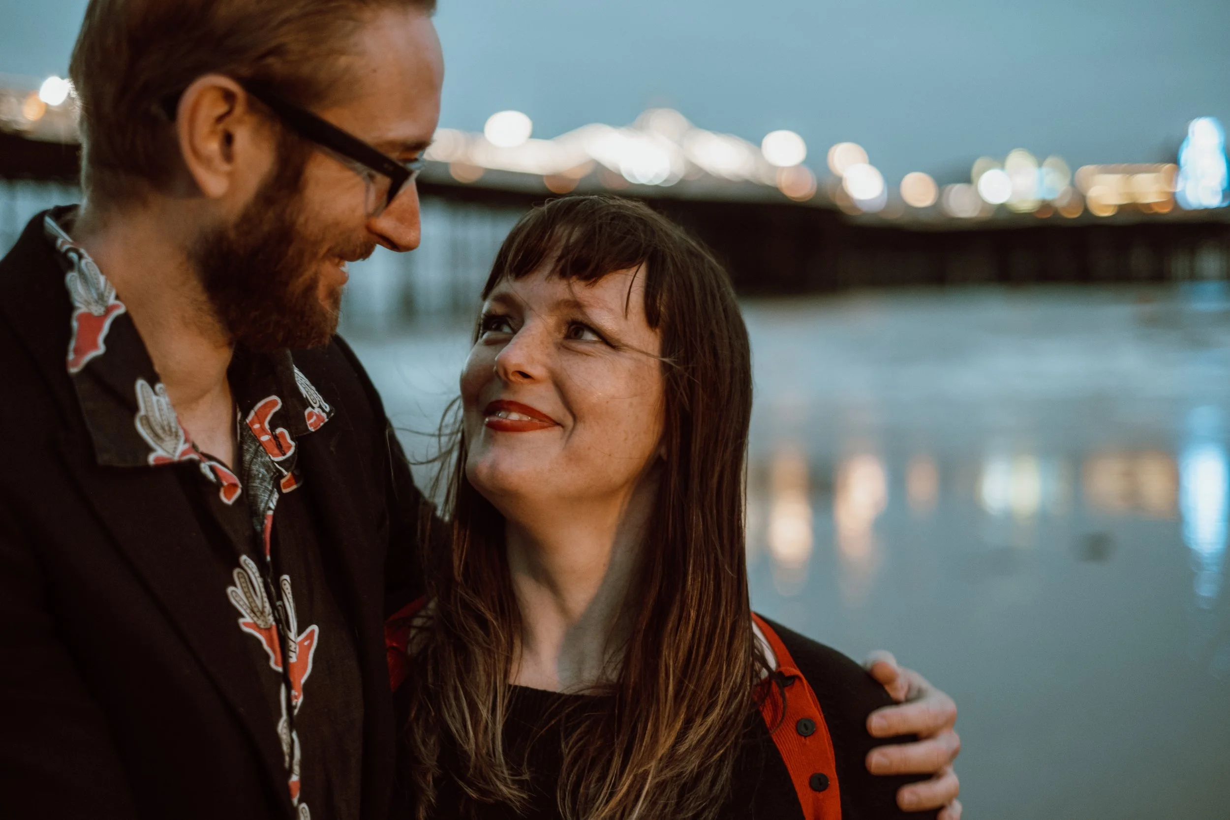 A man and woman looking at each other with a blurred cityscape and water in the background during dusk.