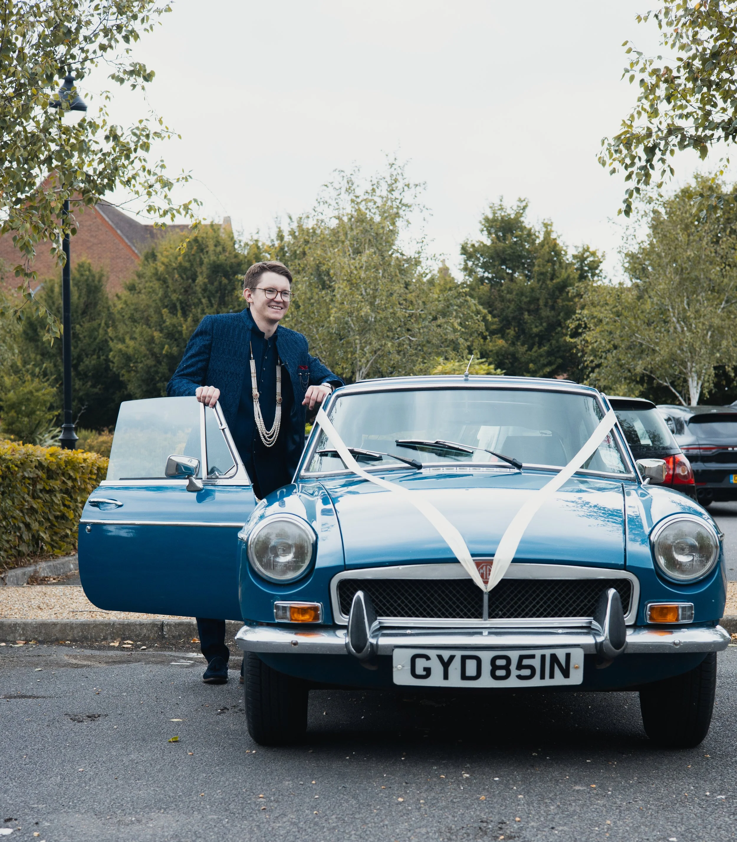 A man in a dark blue jacket and black pants stands next to a vintage blue car decorated with white ribbons, parked in an outdoor lot with trees and other cars in the background.