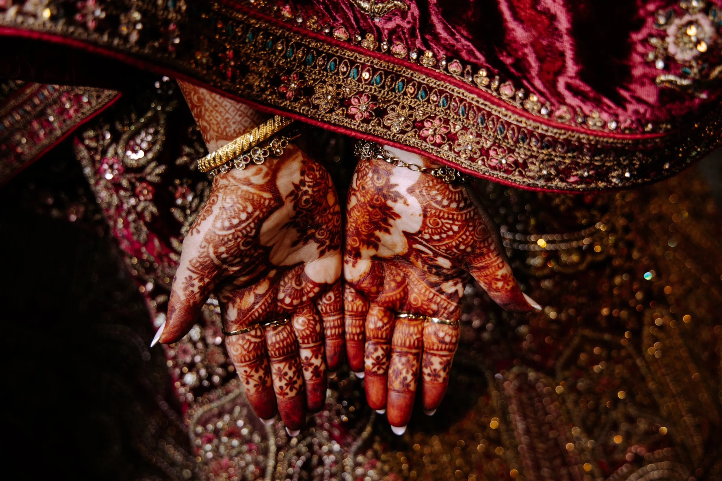 Close-up of hands with intricate henna designs, adorned with rings, jewelry, and highlighted with traditional colorful embroidered attire.