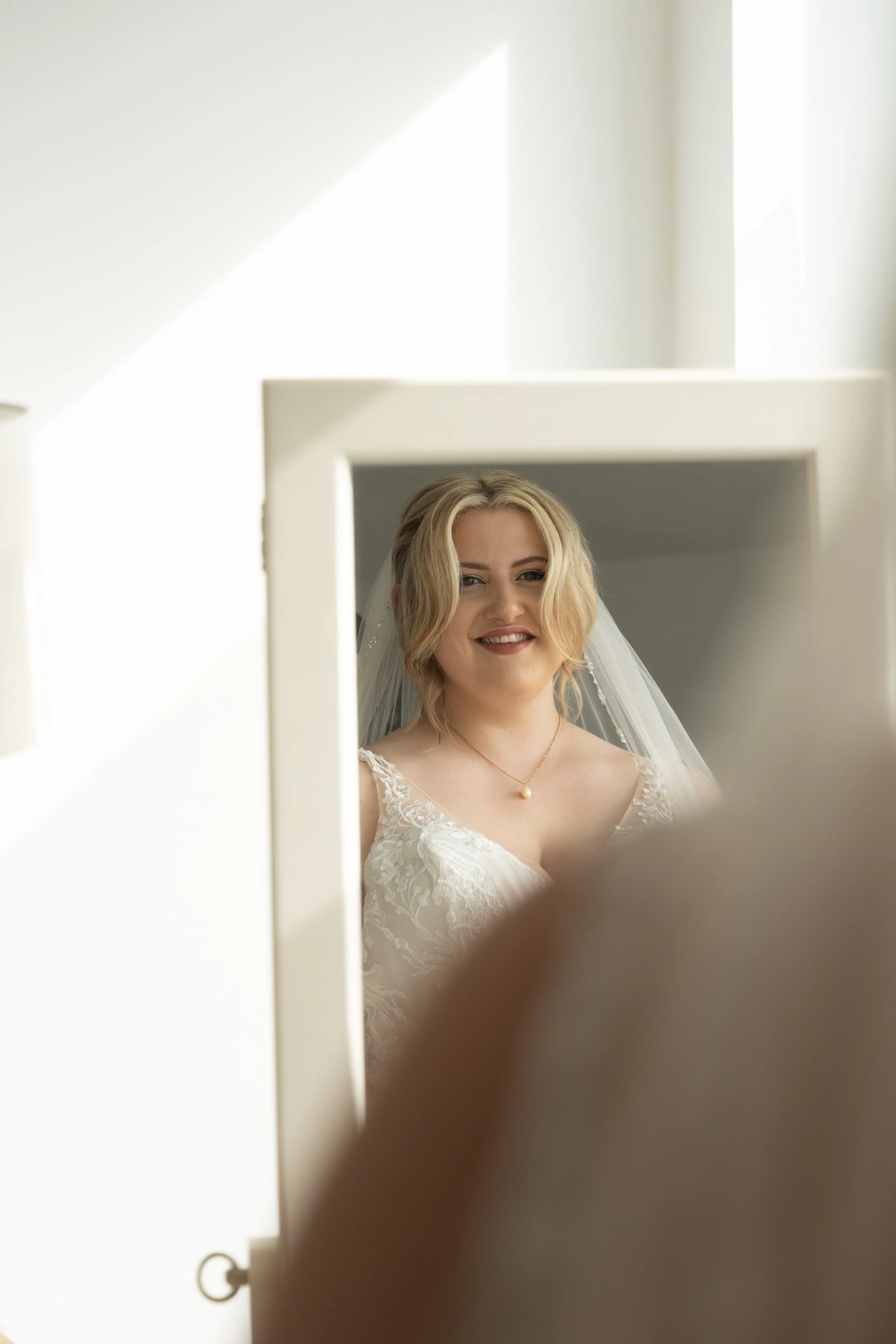 A bride with blond hair styled in loose waves, wearing a white lace wedding dress and a veil, looks at herself in a mirror with a slight smile.